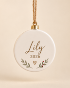 Personalized round ceramic Christmas ornament with name and year on a plain cream background