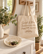 Personalized canvas tote bag reading Sarah & Mia hanging on an entryway hook above a ceramic keepsake dish reading Happy 60th Birthday, Mom holding keys and sunglasses