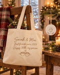 Personalized canvas tote bag reading Sarah & Mia hung on a wooden chair beside a round ceramic ornament reading Lily 2026 in a cozy Christmas room
